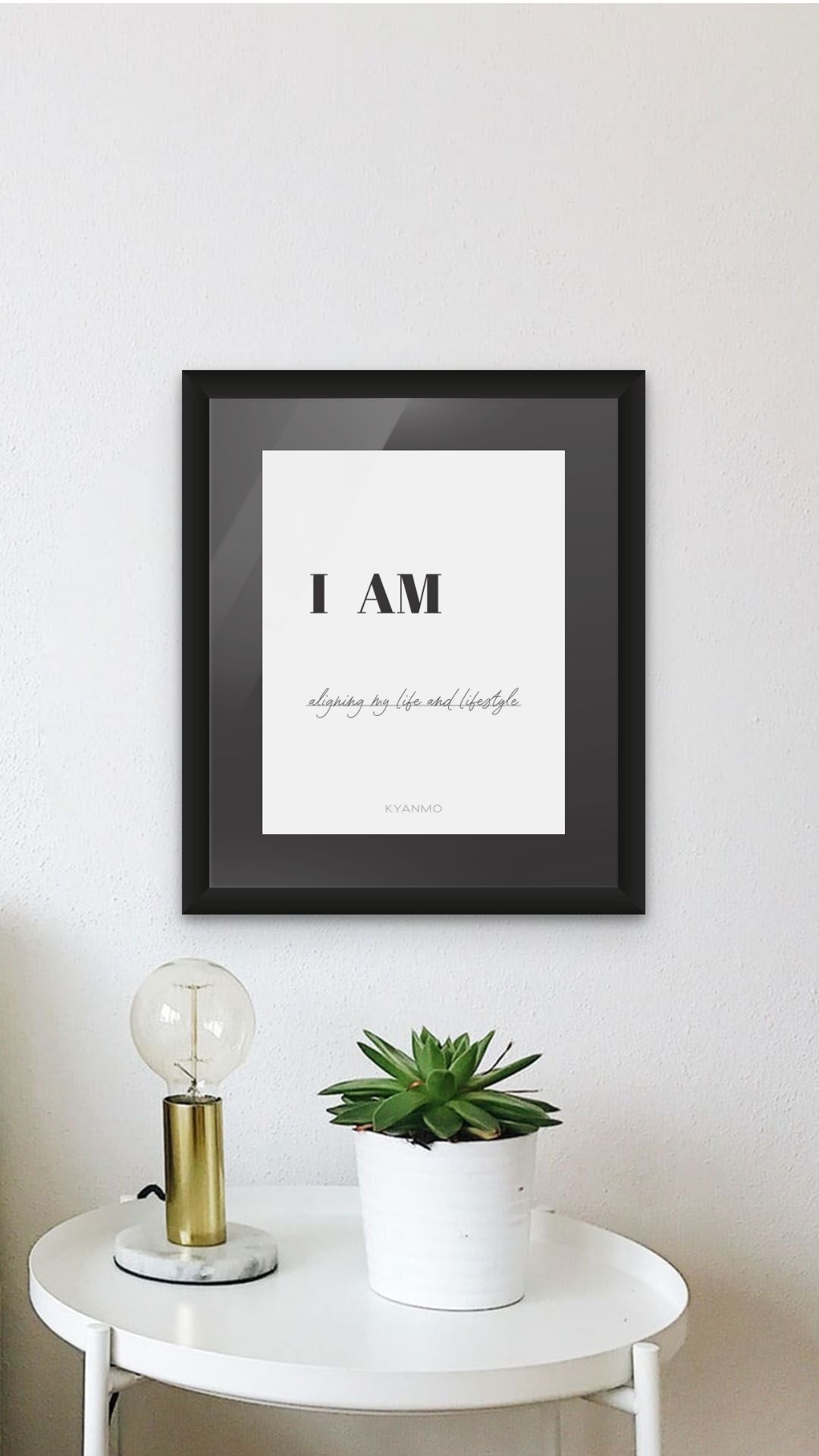 A framed typographic wall art print reading 'I AM' with smaller script text below, displayed on a white wall above a small white side table with a plant and a lamp.