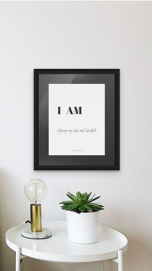 A framed typographic wall art print reading 'I AM' with smaller script text below, displayed on a white wall above a small white side table with a plant and a lamp.